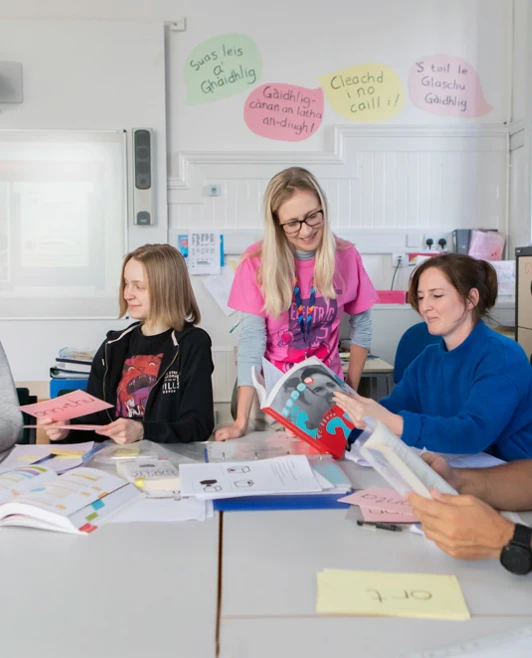 Gaelic language class with students and a lecturer discussing learning materials, surrounded by books, notes, and language flashcards. Gaelic language class with students and a lecturer discussing learning materials, surrounded by books, notes, and language flashcards.